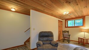A room with a wooden ceiling, a brown leather recliner, a wooden rocking chair, a small table with a lamp, and a window with a view of trees. - Chickasaw in Hocking Hills (Logan)