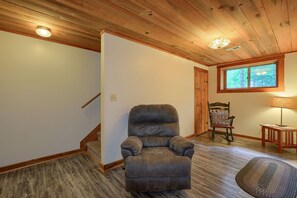 A room with a wooden ceiling, a brown leather recliner, a wooden rocking chair, a small table with a lamp, and a window with a view of trees.