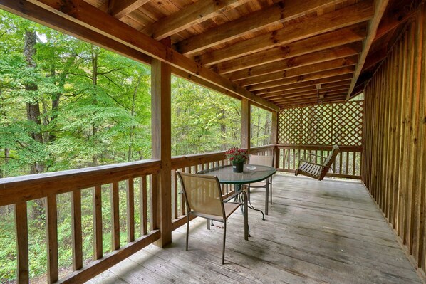 A wooden deck with a table and chairs, surrounded by a wooden railing, overlooking a forested area.