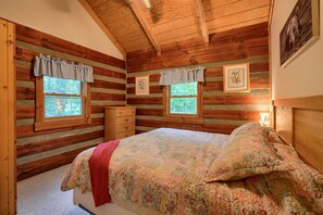Un dormitorio tipo cabaña de madera con dos ventanas, una cama con ropa de cama floral, un aparador y cuadros enmarcados en las paredes.