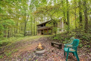 Eine Hütte mit einer Terrasse, umgeben von einem Wald. In der Nähe befindet sich eine Feuerstelle mit Sitzgelegenheiten.