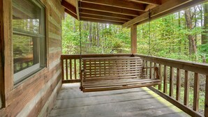 A wooden porch with a bench, a window, and a view of a forest. - Cherokee in Hocking Hills (Logan)