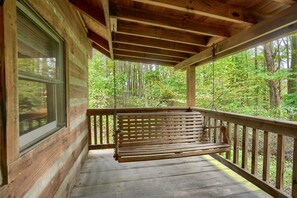 A wooden porch with a bench, a window, and a view of a forest. - Cherokee in Hocking Hills (Logan)
