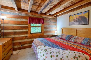 A log cabin bedroom with a wooden ceiling, a large bed with a colorful quilt, a window with a red valance, and a bear painting on the wall. - Cherokee in Hocking Hills (Logan)