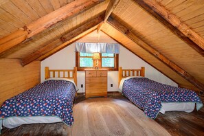 Une chambre mansardée chaleureuse, avec deux lits, une commode en bois et une fenêtre donnant sur les arbres.