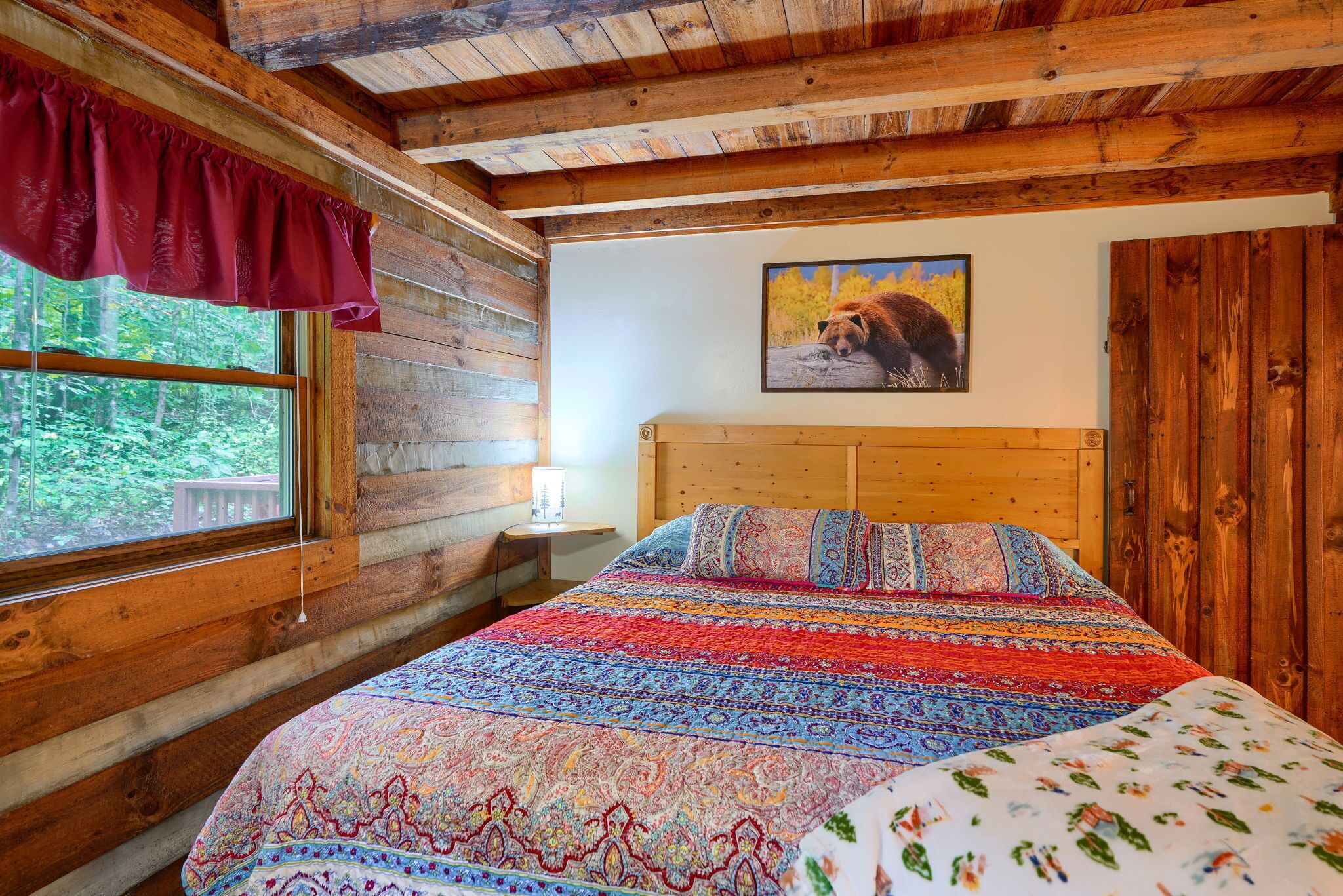 Un dormitorio de cabaña de madera con una cama grande, una ventana con vista a árboles, un cabecero de madera y un cuadro de un oso en la pared.