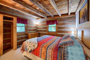 A log cabin bedroom with a large bed, two windows with curtains, a wooden dresser, and a closet. - Cherokee in Hocking Hills (Logan)