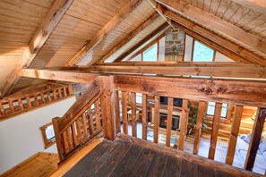 A wooden staircase with a railing leading to a lofted area with a stone fireplace and a view of the outdoors.