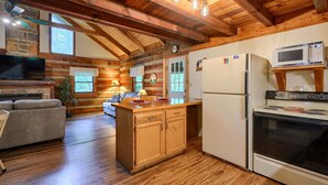 A log cabin interior with a wooden dining table, a built-in kitchen with a stove and microwave, a sofa, and a stone fireplace. - Cherokee in Hocking Hills (Logan)