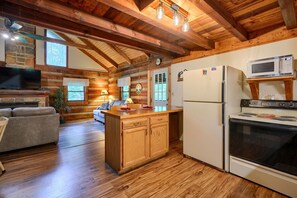 A log cabin interior with a wooden dining table, a built-in kitchen with a stove and microwave, a sofa, and a stone fireplace.