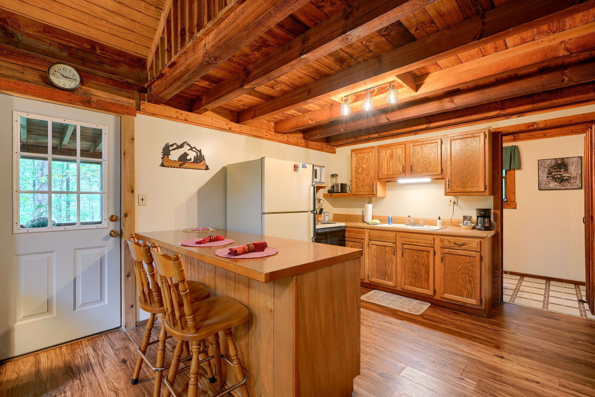A wooden cabin interior with a kitchen area featuring wooden cabinets, a refrigerator, and a bar counter with stools.