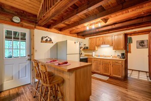 A wooden cabin interior with a kitchen area featuring wooden cabinets, a refrigerator, and a bar counter with stools.