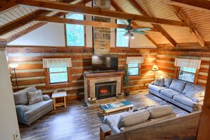 A log cabin interior with a fireplace, two sofas, a television, and a ceiling fan.
