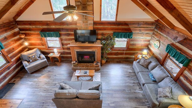 Interior de una cabaña de troncos con chimenea, dos sofás, un televisor y un ventilador de techo.