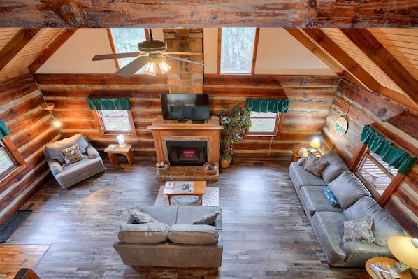 A log cabin interior with a fireplace, two sofas, a television, and a ceiling fan.