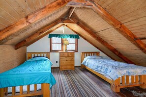 Une petite chambre mansardée avec deux lits en bois, une commode en bois et une fenêtre avec un rideau.