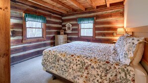 Un dormitorio tipo cabaña de madera con una cama de madera, dos ventanas con cortinas, un tocador y una mesita de noche con una lámpara.