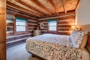 Une chambre dans un chalet en rondins, avec un lit en bois, deux fenêtres avec des rideaux, une commode et une table de chevet avec une lampe.