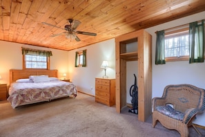 A bedroom with a wooden ceiling, a bed with a floral bedspread, a wooden dresser, a wicker chair, and a window with curtains.