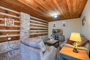 A cozy living room with a stone wall, wooden ceiling, and a fireplace. There is a sofa, a coffee table, and a lamp. - Navajo in Hocking Hills (Logan)