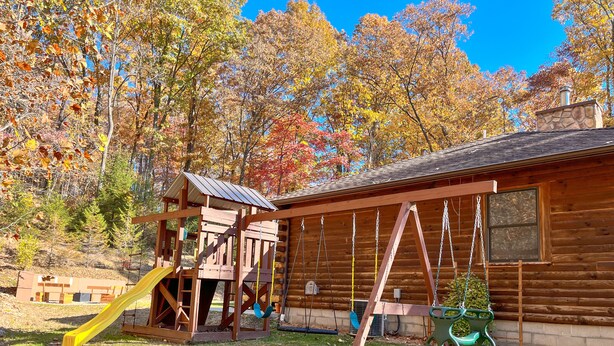 A wooden cabin with a swing set and a slide in a grassy area surrounded by trees with autumn foliage.