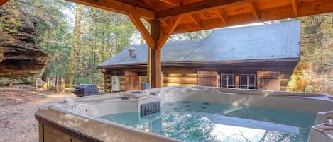A hot tub under a wooden gazebo with a stone wall and a cabin in the background.