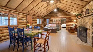 En stuga byggd av timmer med en inredning som inkluderar en matplats, en soffa, en öppen spis och en takfläkt.