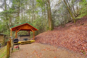 Una estructura de madera rústica con techo verde, una parrilla para barbacoa y una mesa de picnic en un área boscosa con hojas caídas.