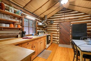 A rustic cabin interior with wooden walls, a kitchen with wooden cabinets, a dining table, and a black refrigerator.