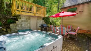 A hot tub with a stone exterior, surrounded by a patio with striped chairs and a red umbrella. - Hillside Hideaway in Hocking Hills (Logan)