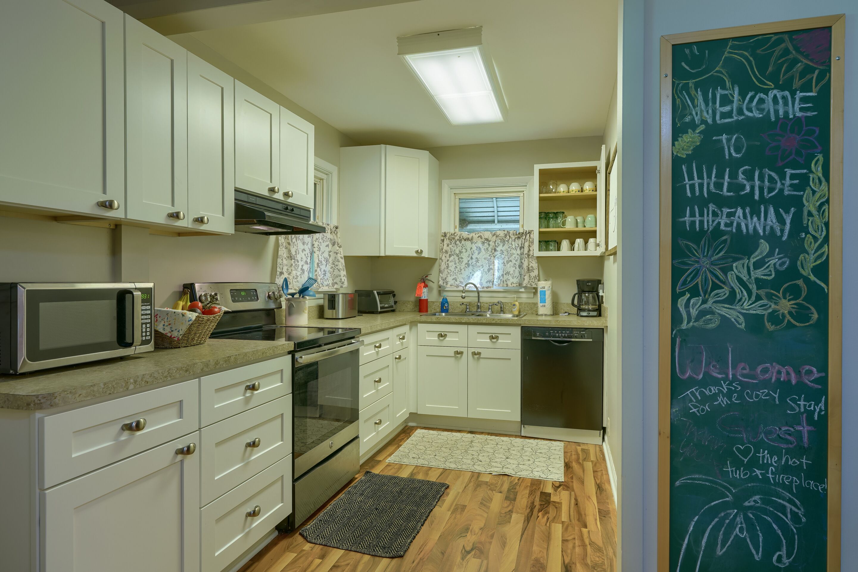 A modern kitchen with white cabinets, a microwave, a toaster, and a chalkboard with a welcome message.