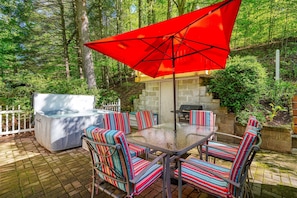 An outdoor patio with a glass-top table, striped chairs, a red umbrella, a hot tub, and a barbecue grill. - Hillside Hideaway in Hocking Hills (Logan)