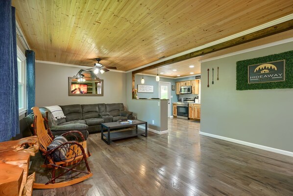 A spacious living area with a wooden ceiling, a green sofa, a rocking chair, and a kitchen in the background. - Hideaway Pines Cabin (Logan)