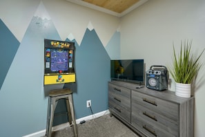 Ein Zimmer mit einem Pac-Man-Spielgerät, einem Fernseher, einem Radio, einer Pflanze und einem Holzschemel.
