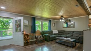 A spacious living room with a wooden ceiling, a green sofa, a wooden coffee table, and a view of the outdoors through large windows. - Hideaway Pines Cabin (Logan)