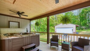 A covered outdoor kitchen area with a built-in stove, a ceiling fan, and a view of a garage and trees. - Hideaway Pines Cabin (Logan)