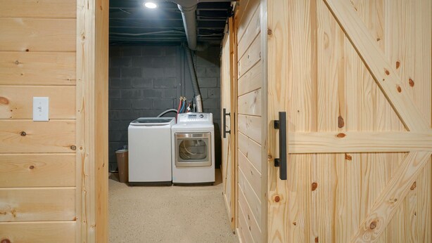 A laundry room with two washing machines and a dryer, wooden walls, and a concrete floor. - Hideaway Pines Cabin (Logan)