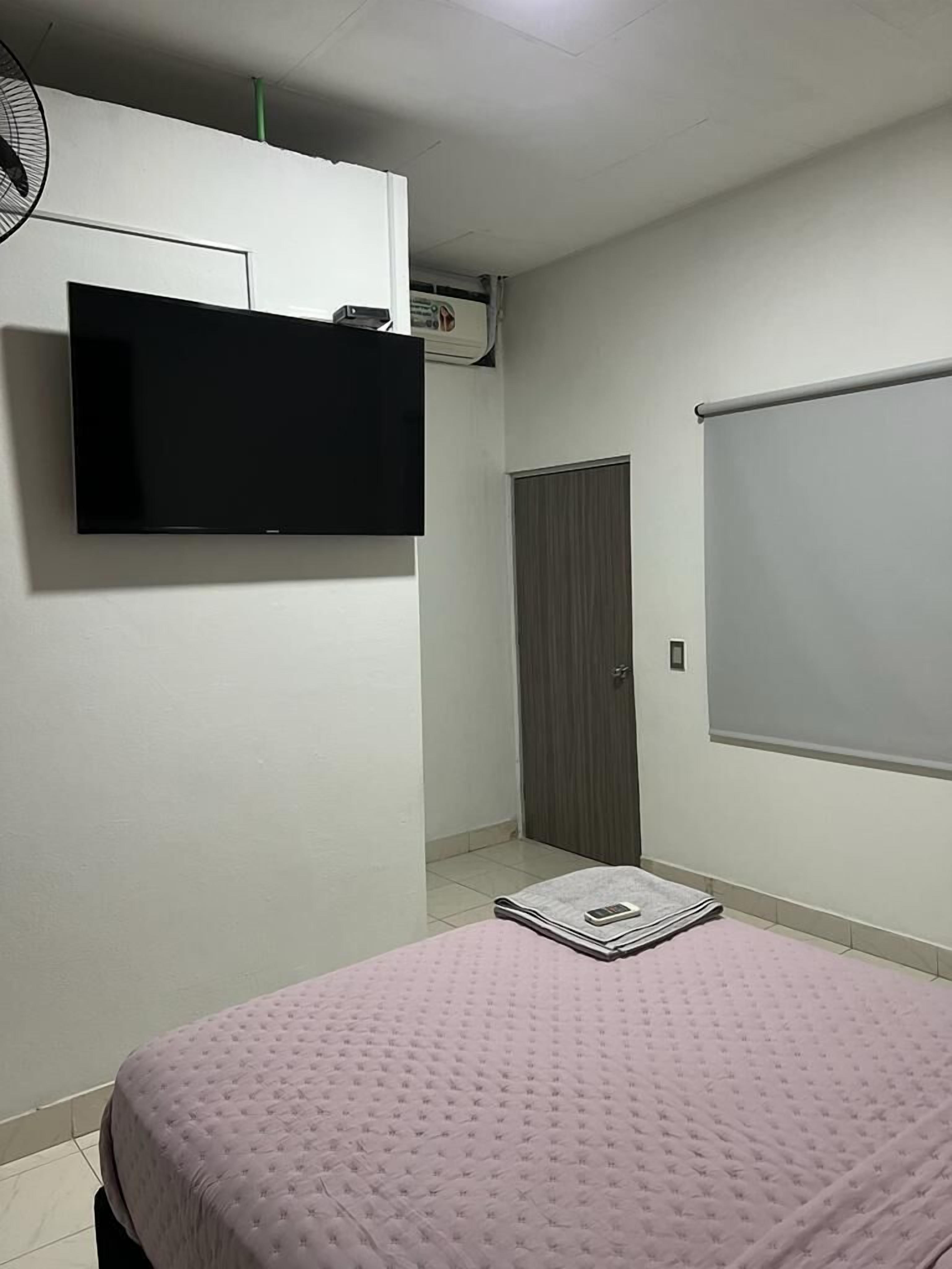 A bedroom with a bed, a flat-screen TV mounted on the wall, a ceiling fan, and a window with blinds.