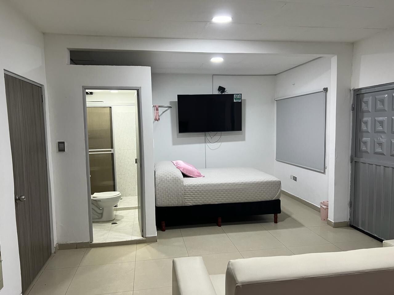 Una habitación pequeña y bien iluminada con una cama, un televisor montado en la pared y una puerta que da a un baño.