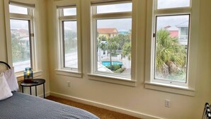 Un dormitorio con una cama grande, una mesita de noche pequeña con lámpara y tres ventanas que dan a una piscina y palmeras.