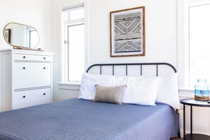 A bed with a blue bedspread, a white pillow, and a beige pillow. There is a white dresser with drawers and a round mirror on top. A framed artwork hangs on the wall above the bed.