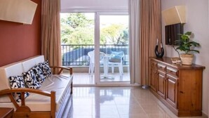 Ein Zimmer mit einer Couch, einem Holzschrank, einem Fernseher und einem Balkon mit Tisch und Stühlen.