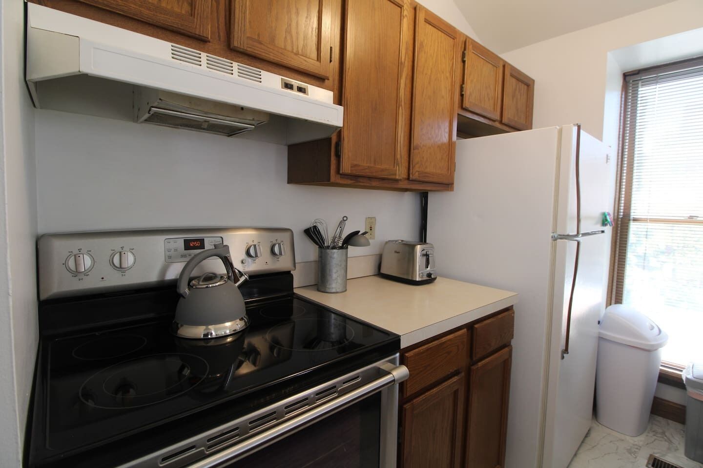 A compact kitchen with wooden cabinets, a stove, a kettle, a toaster, and a refrigerator.