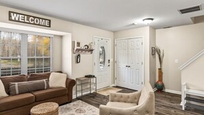 Ein Wohnzimmer mit einem braunen Sofa, einem beige farbenen Sessel, einem 'WELCOME'-Schild, einer weißen Tür und einer Vase mit Zweigen.