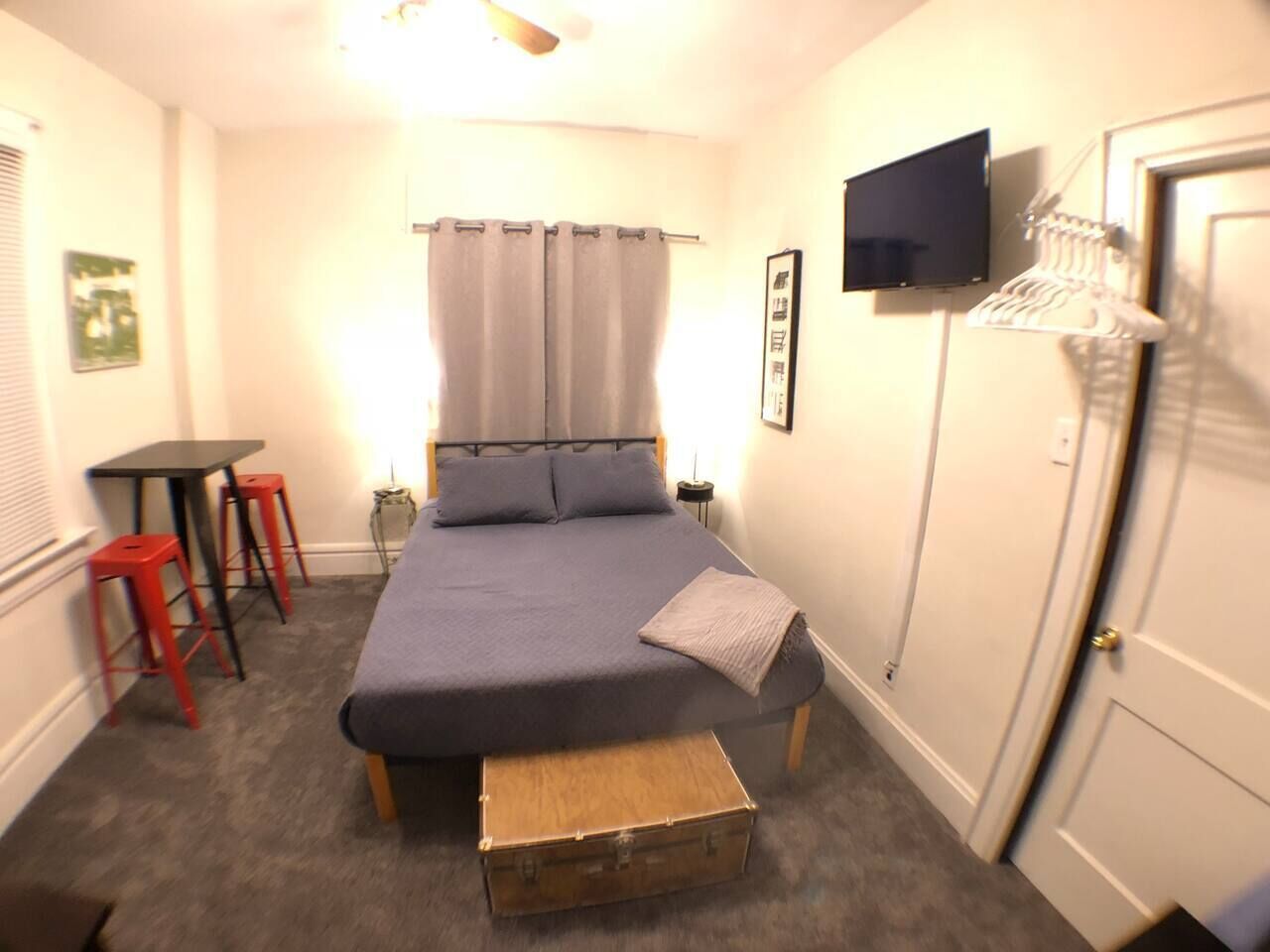 Ein Schlafzimmer mit einem Bett, einem kleinen Tisch, zwei roten Hocker, einem Fernseher an der Wand und einer Tür.