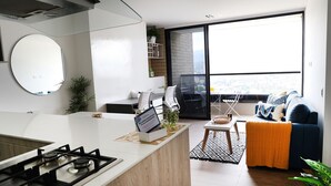 Una cocina moderna con estufa a gas, un comedor con mesa y sillas blancas, y una sala de estar con sofá azul y un amplio ventanal con vista a la ciudad.