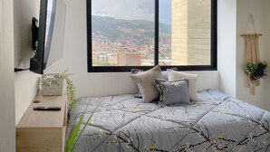 Un dormitorio con cama, televisión, ventana con vista a la ciudad, escritorio de madera y un colgador de macramé en la pared.