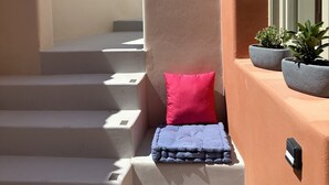 Un espace extérieur ensoleillé, agrémenté d’un escalier beige menant à un petit coin salon avec un coussin rouge et une couverture bleue. Deux plantes en pot se trouvent sur le côté droit.