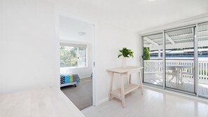 A bright room with a white wall, a wooden table with a plant, and a view of a balcony with a white fence. - Whalesong - by Lifestyle. Brunswick Heads (NEW BRIGHTON)
