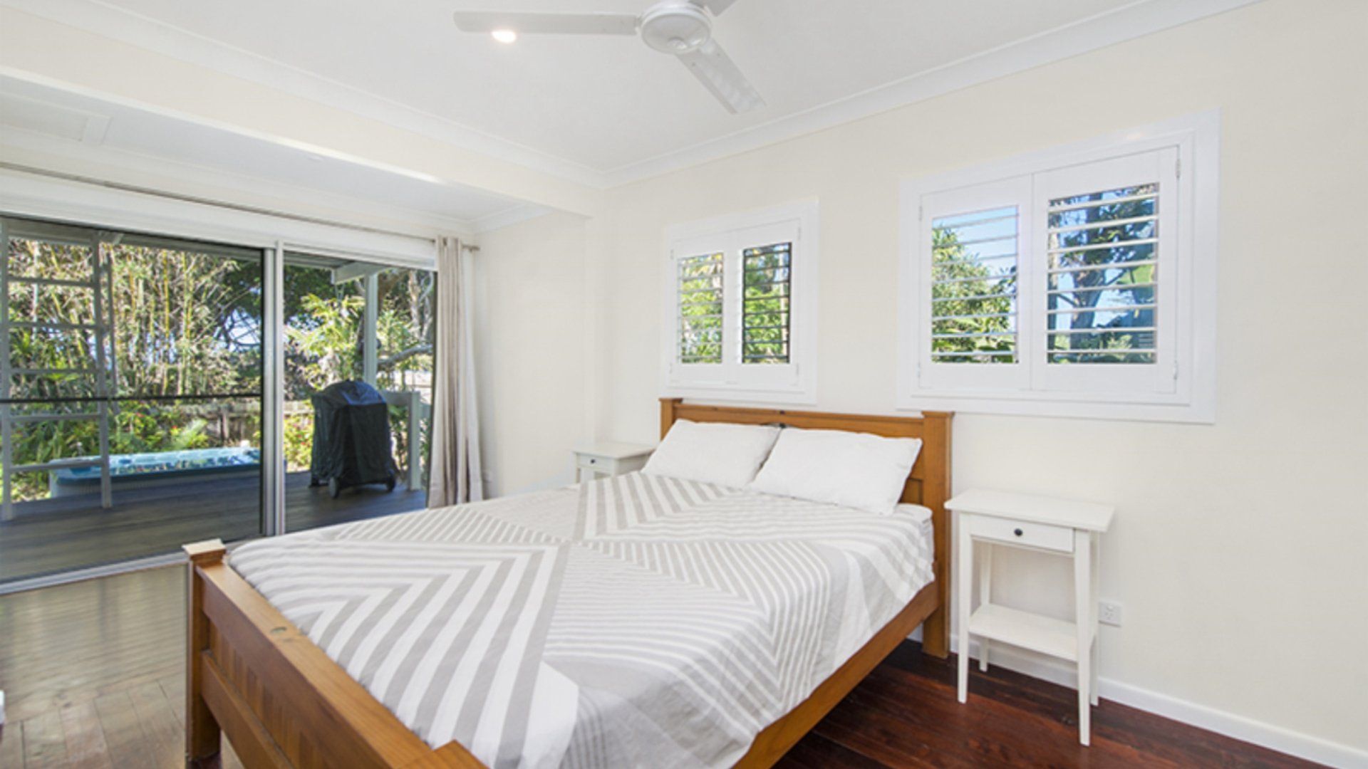 Un dormitorio con cama, mesita de noche y ventilador de techo. Hay ventanas con persianas y vistas al exterior con zonas verdes.